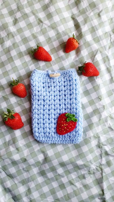 A kindle sleeve that is blue and has a crocheted strawberry sewn onto the front